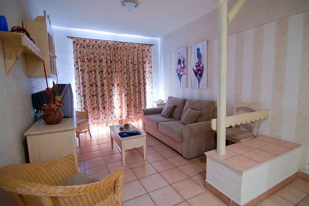 Un salon comprenant un canapé, une table basse, un fauteuil et un téléviseur. Il y a une fenêtre avec des rideaux et un mur orné de tableaux.