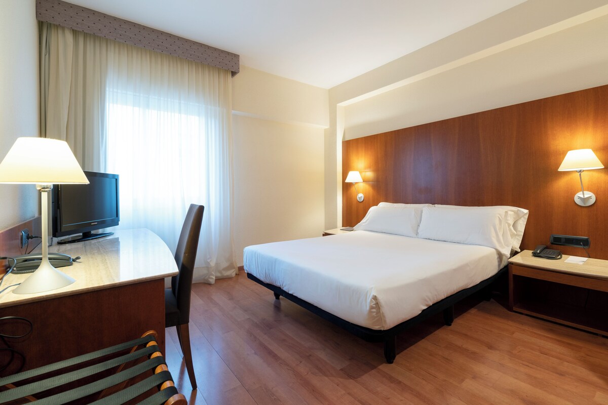 A hotel room with a bed, desk, chair, TV, and two bedside lamps.