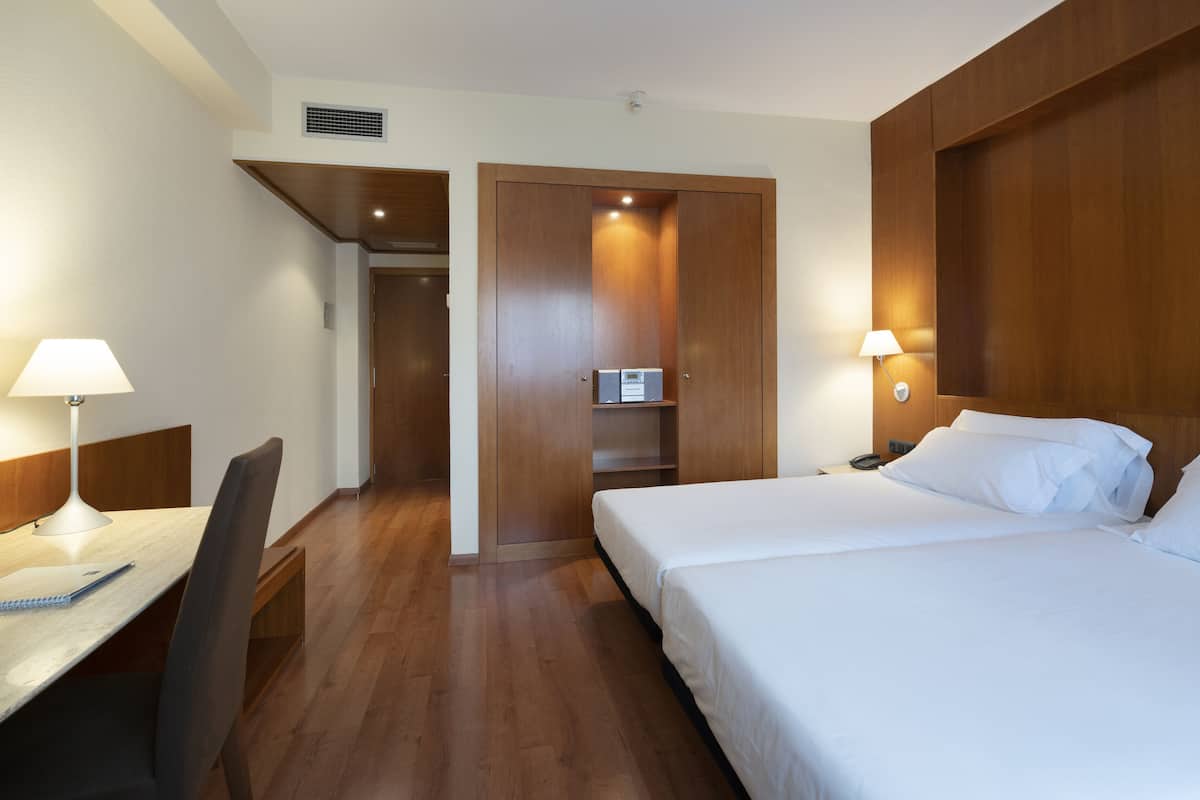 Een hotelkamer met een houten vloer, een bureau met lamp, een bed met witte lakens en een kledingkast met spiegel.
