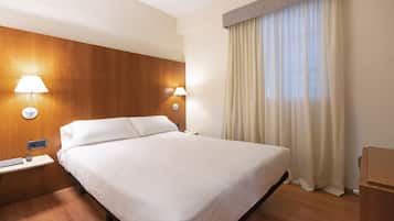 Habitación de hotel con una cama grande, dos lámparas de noche, cabecero de madera, mesita de noche y una ventana con cortinas.