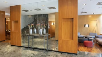 A modern lobby with a glass partition, wooden panels, a marble floor, and a seating area with sofas and a red ottoman.
