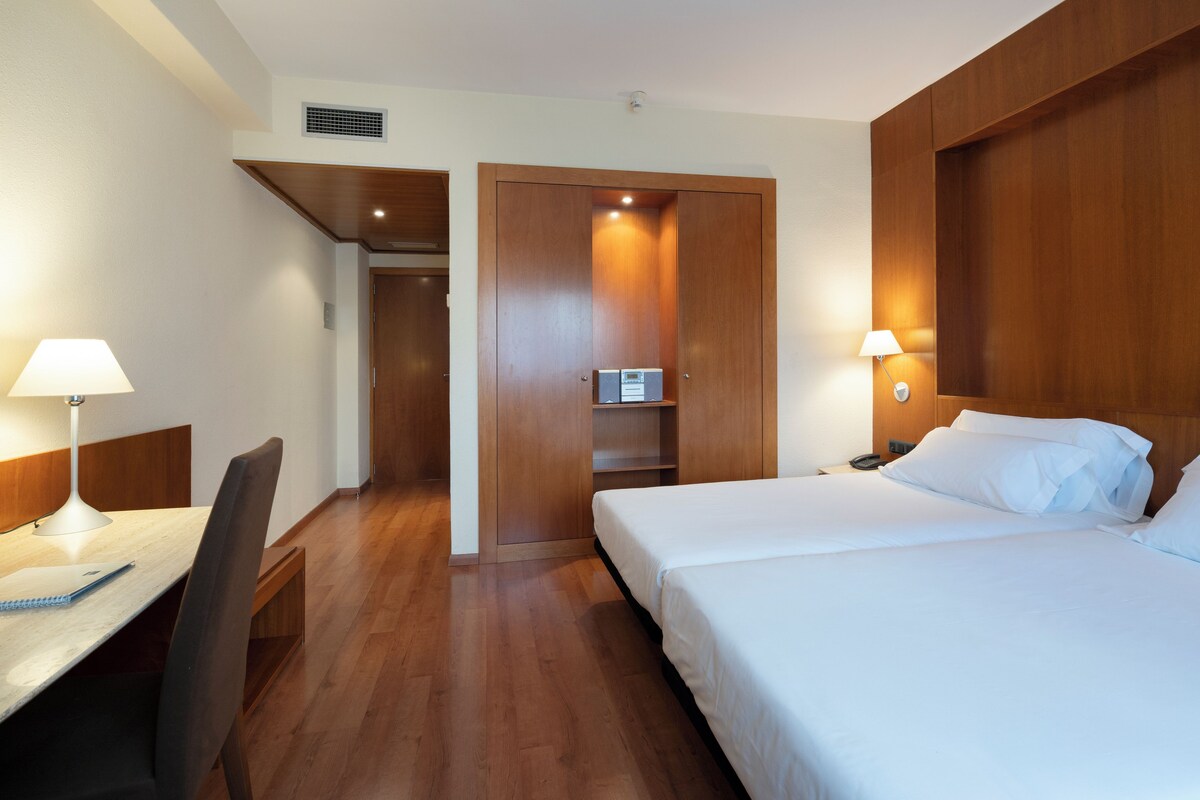 Quarto de hotel com escrivaninha de madeira, duas camas e um guarda-roupa.