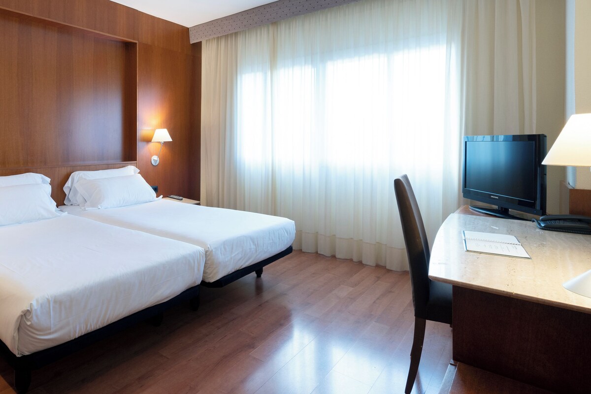 Quarto de hotel com duas camas, uma secretária com televisão e duas luminárias.