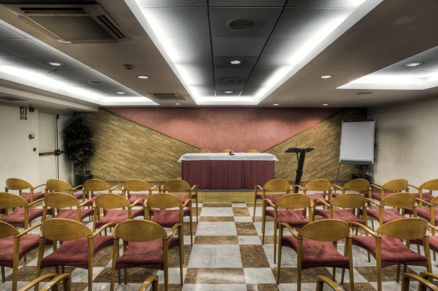 Un salón de conferencias con filas de sillas, un escenario con una pizarra y un panel de pared de madera.