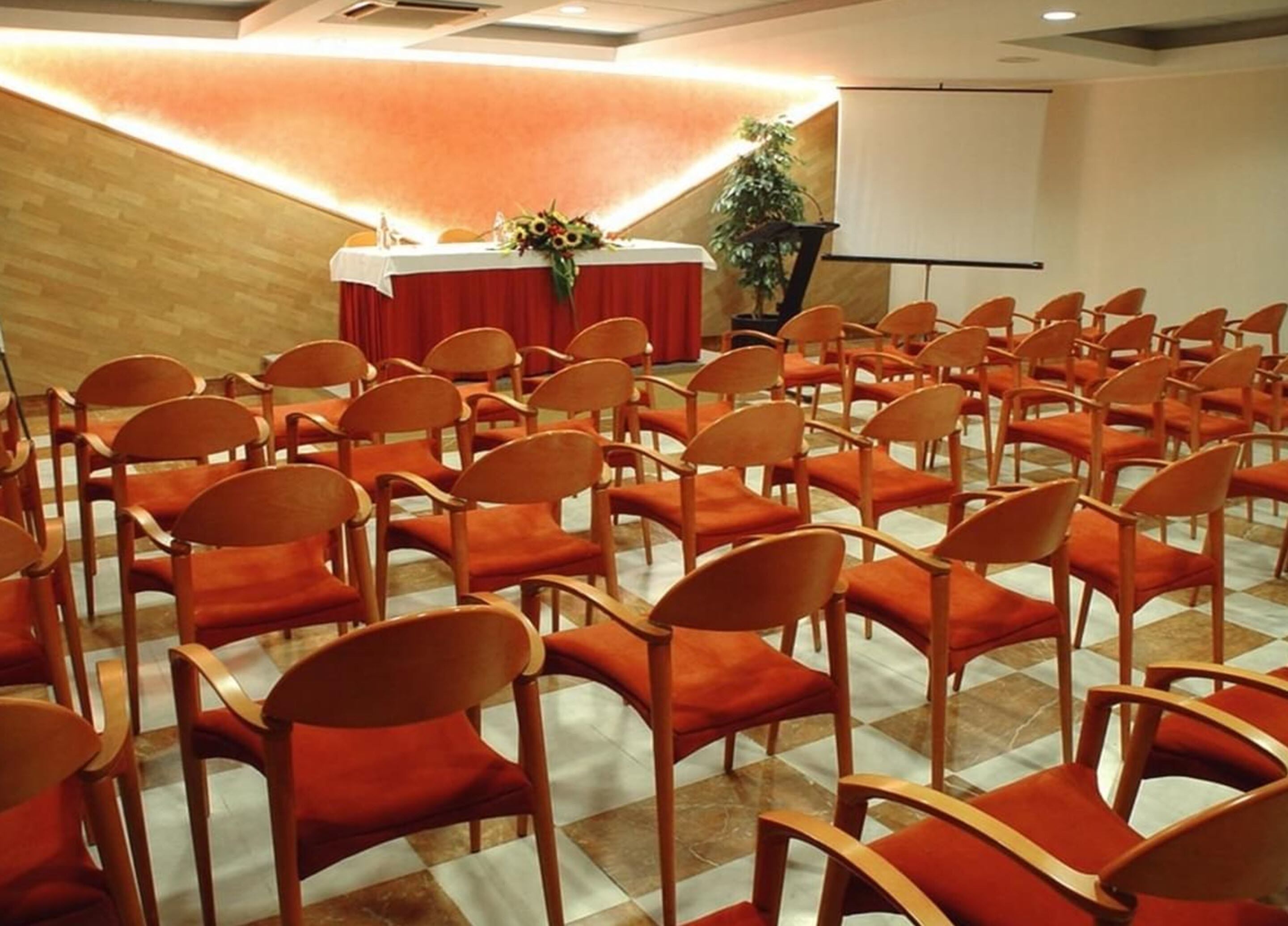 Ein Konferenzraum mit Reihen orangener Stühle, einem Tisch mit rotem Tischlein und einer Projektionsleinwand.