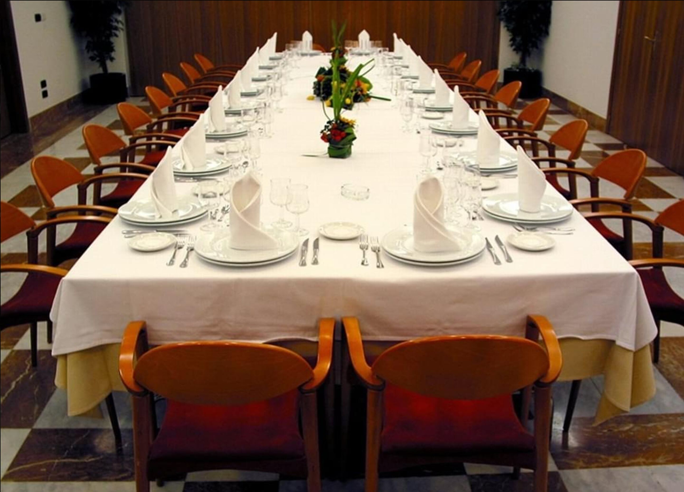A long banquet table set with white tablecloths, silverware, and glassware, surrounded by orange chairs.