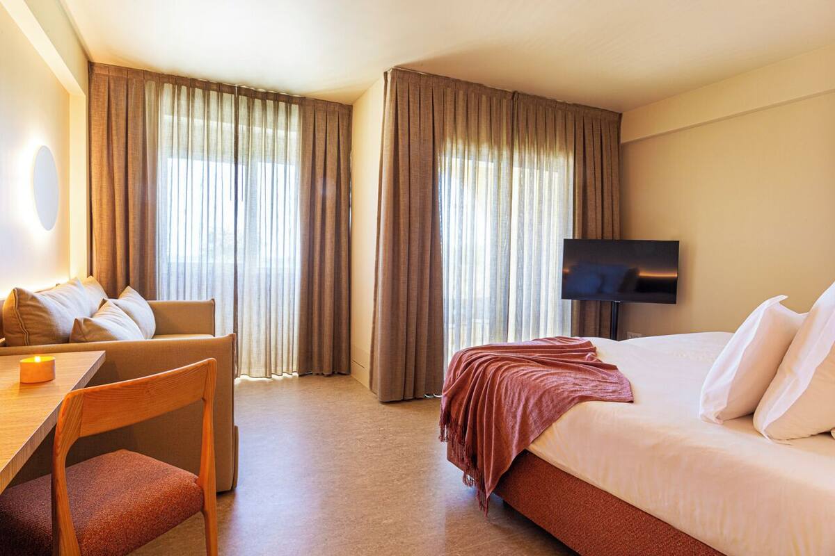 A hotel room with a bed, a desk with a chair, a TV, and large windows with curtains.
