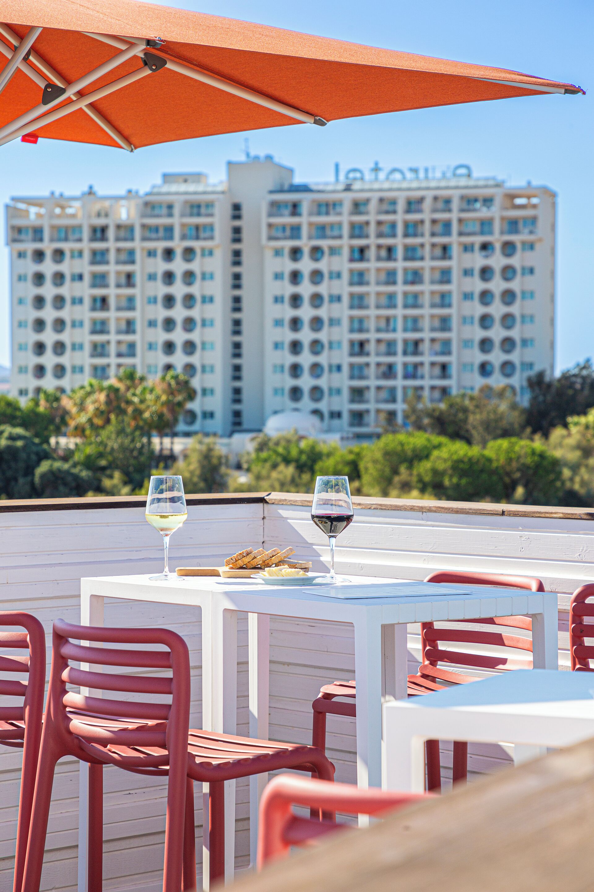 Una terrazza con tavoli bianchi e sedie rosse, due bicchieri di vino e vista su un edificio di un hotel sullo sfondo.