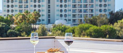 Una terrazza con tavoli bianchi e sedie rosse, due bicchieri di vino e vista su un edificio di un hotel sullo sfondo.