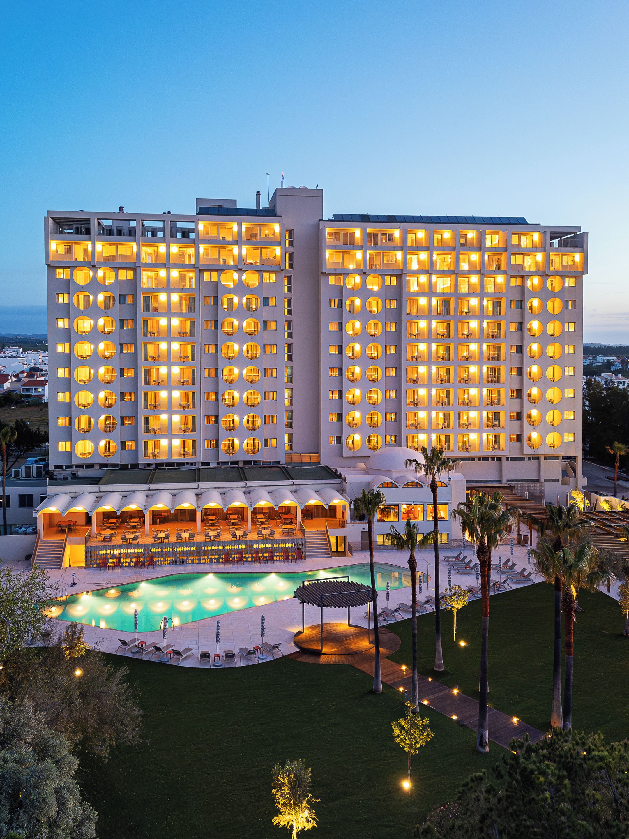 A multi-story hotel with a pool area, outdoor seating, and a well-lit facade.