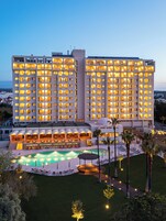 Un hotel de varios pisos con área de piscina, mobiliario exterior e iluminación destacada en la fachada.