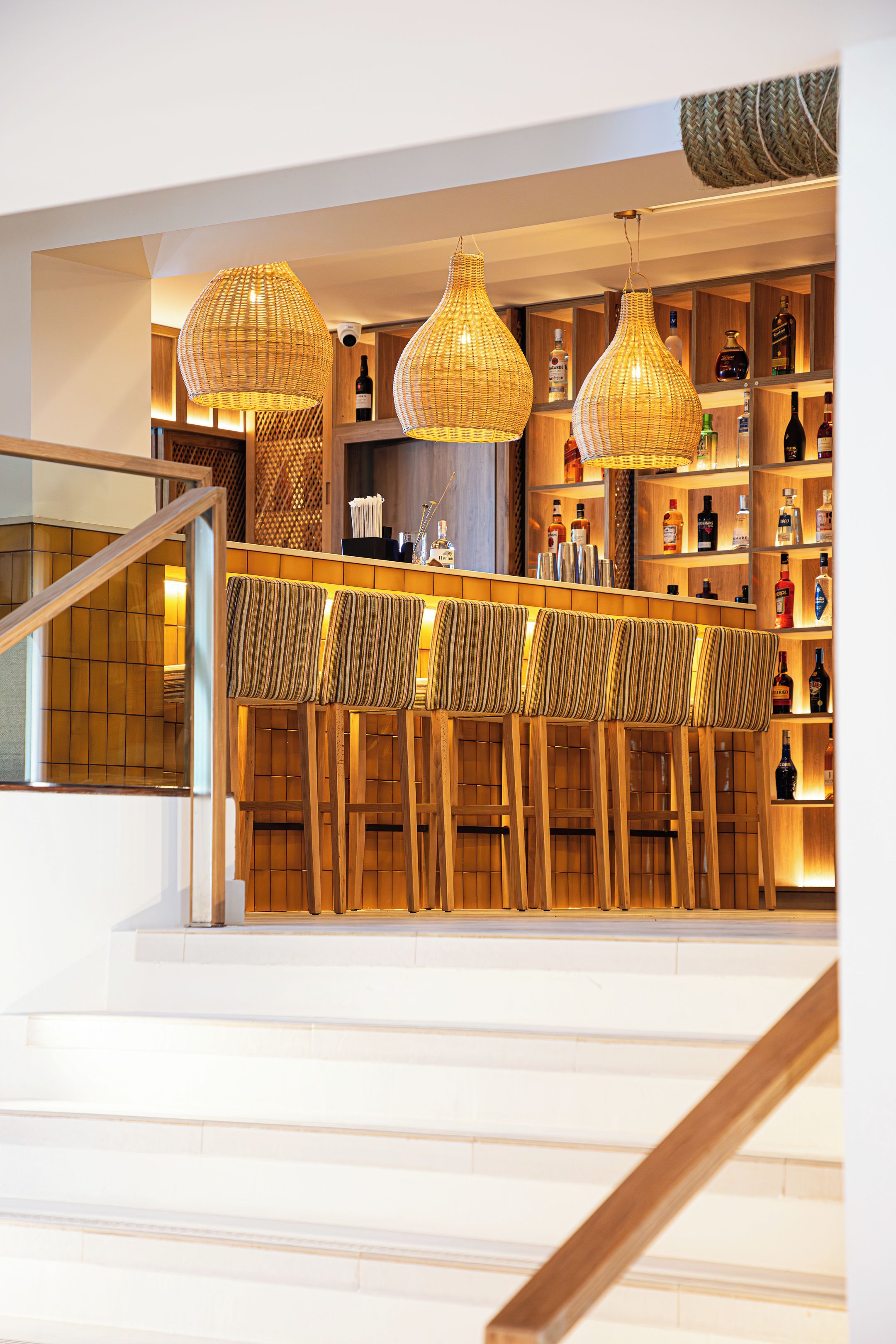 A bar area with wicker pendant lighting, wooden bar stools, and shelves stocked with liquor bottles.