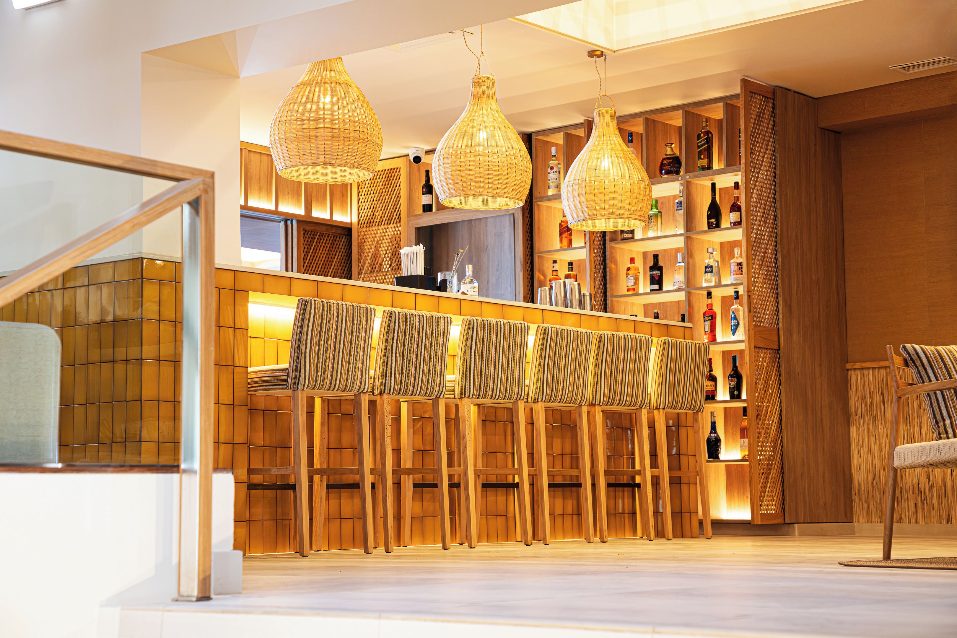 Una zona de bar con mobiliario de madera, lámparas de mimbre colgantes y estanterías llenas de botellas.