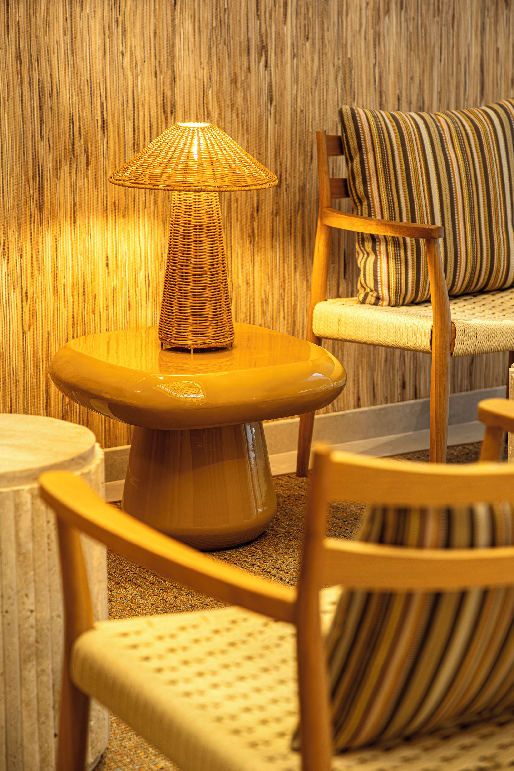 Une lampe en osier posée sur une table ronde, un canapé à rayures et une chaise en bois dans une pièce aux murs recouverts de lambris en bois verticaux.