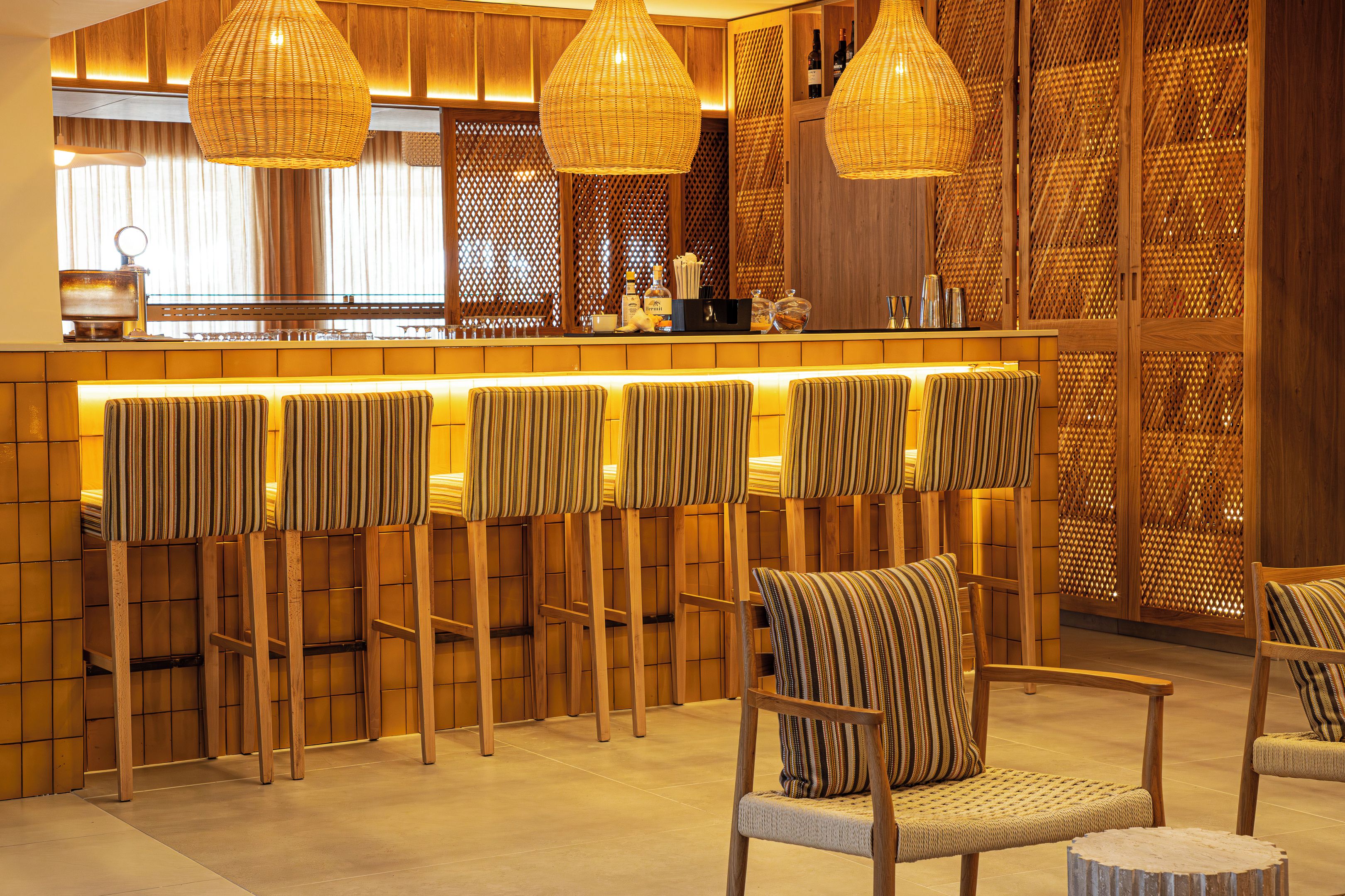 A bar area with multiple seating options, including bar stools and chairs, with a wooden backdrop and hanging pendant lights.