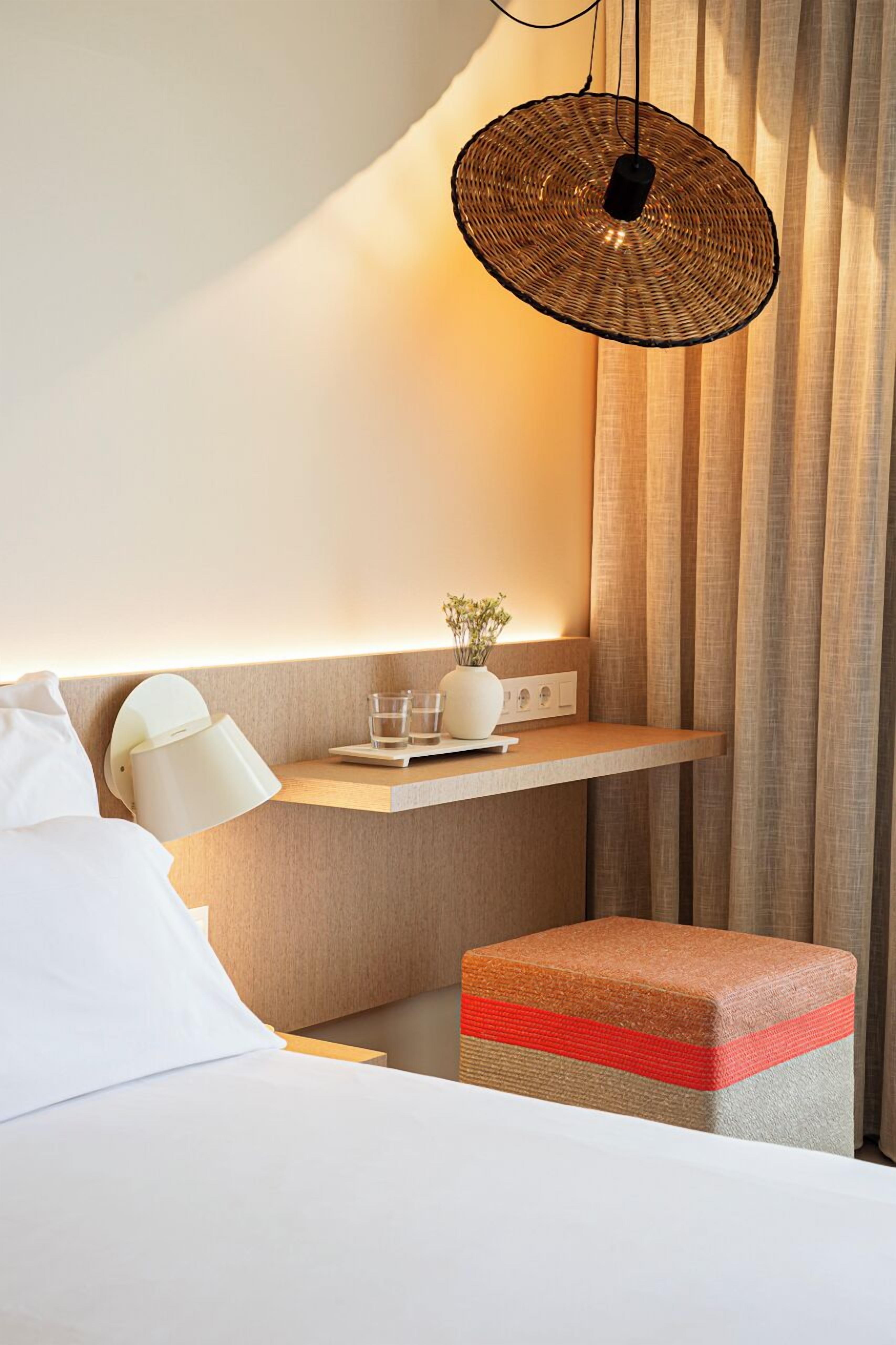 A modern hotel room with a bed, bedside table, lamp, and a small shelf with glasses and a vase.