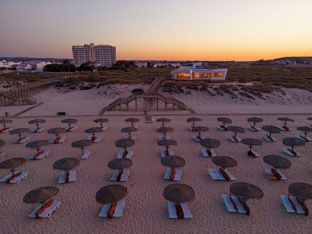 Una playa con filas de tumbonas y sombrillas, un paseo marítimo que lleva a un edificio, y un hotel al fondo.