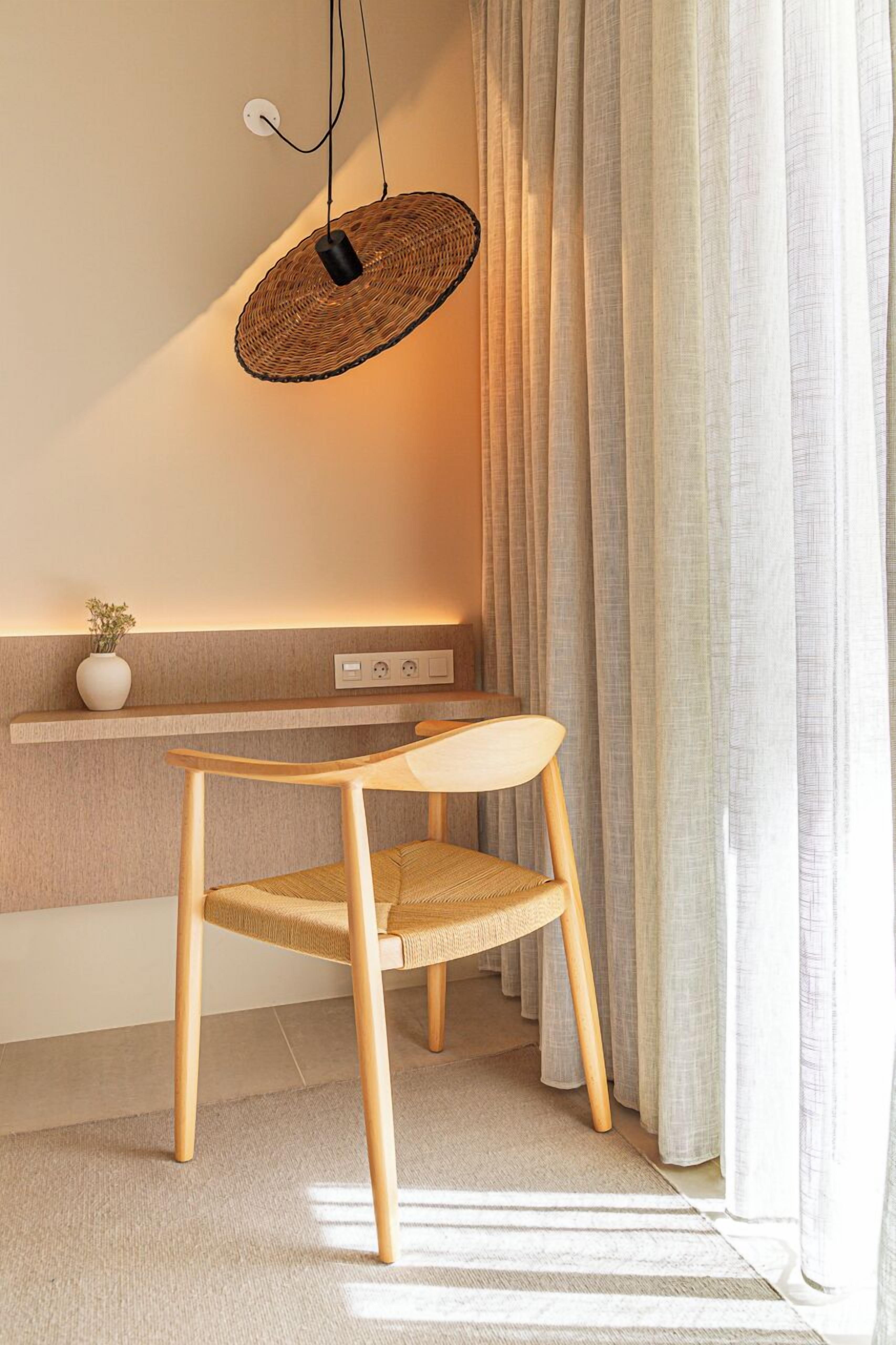 Una habitación moderna con un escritorio de madera, una silla tejida, una lámpara colgante y una ventana con cortinas.