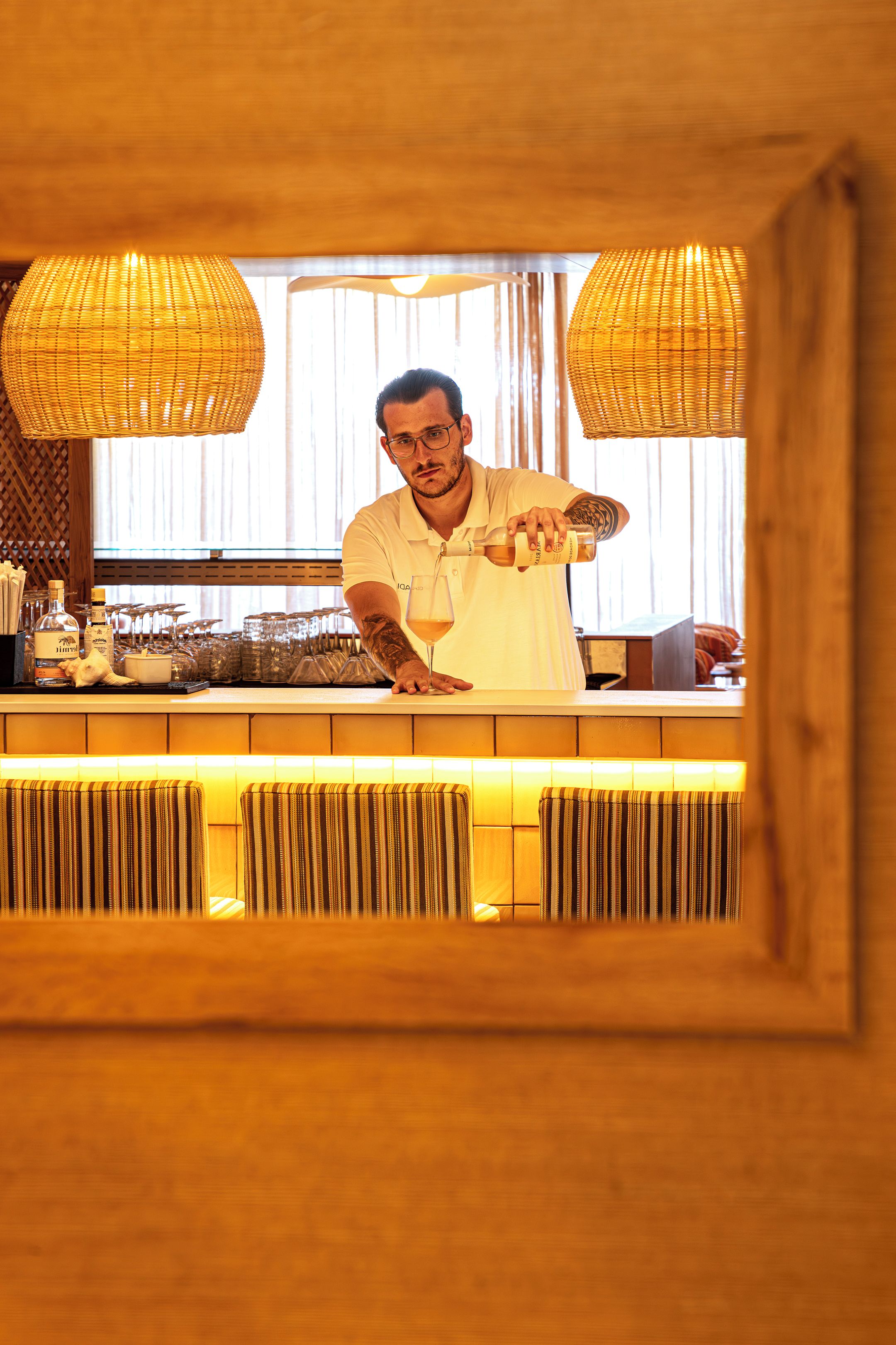 Uma área de bar com um bartender servindo uma bebida, um balcão com copos e garrafas, e uma janela com cortinas.