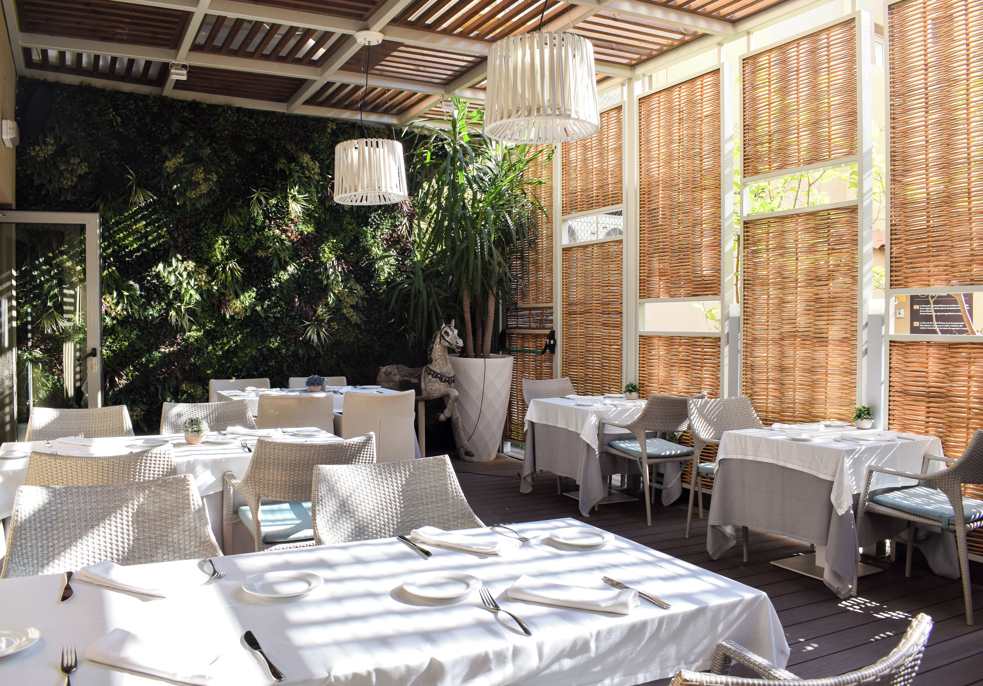 An outdoor dining area with tables set for two, surrounded by wicker furniture and a decorative horse statue.
