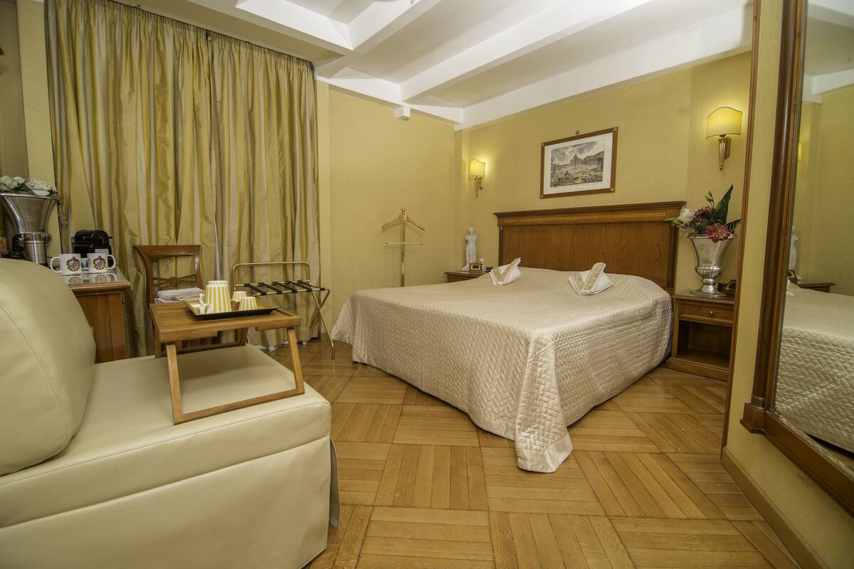 A hotel room with a bed, a chair, a desk with a lamp, a mirror, and a painting on the wall.