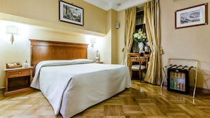 A hotel room with a large bed, bedside tables, a desk with a chair, a refrigerator, and two framed pictures on the walls. - Luxury Rooms H 2000 Roma (Rome)