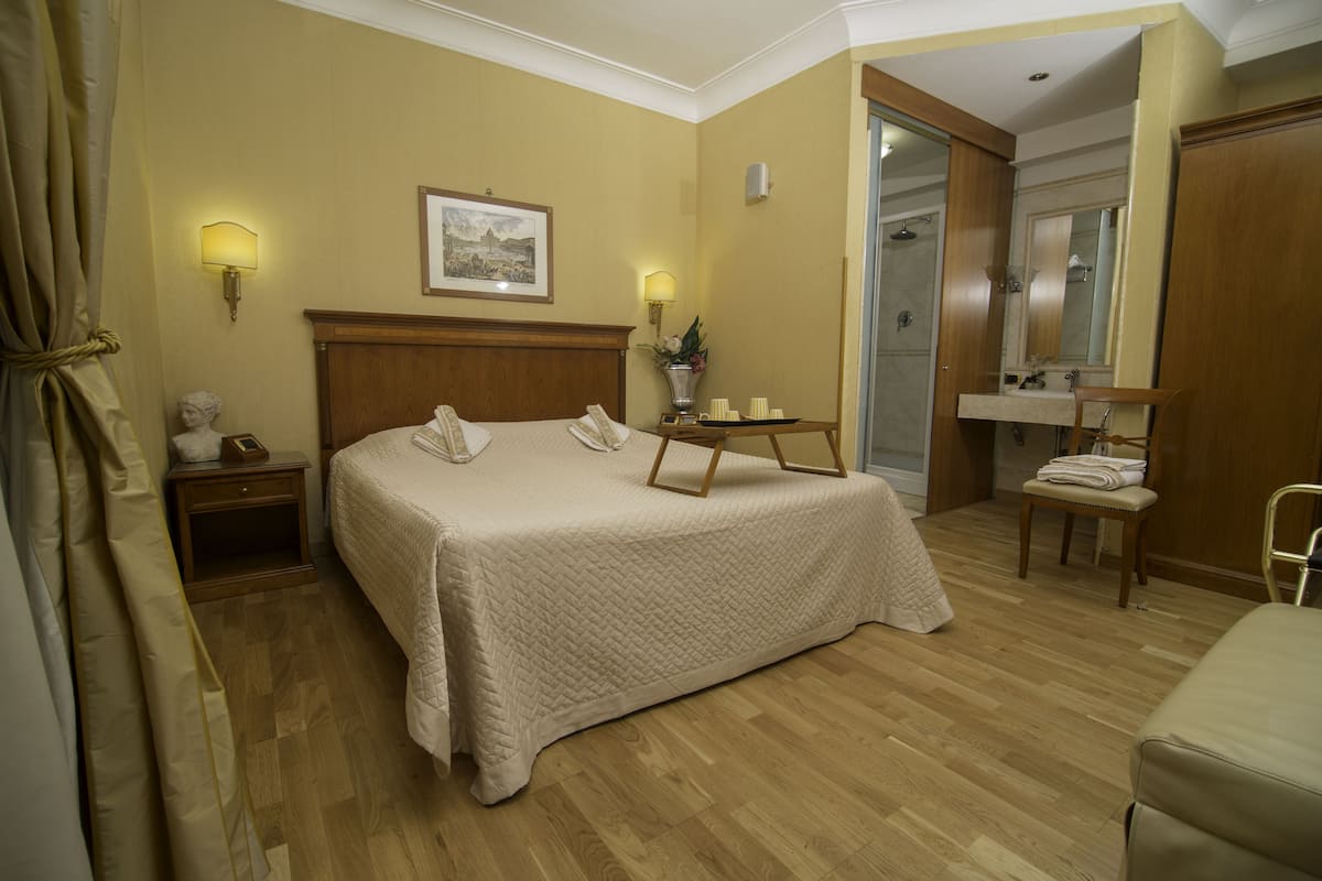 A hotel room with a large bed, bedside lamps, a bedside table with a phone and a vase, a wooden chair, a wooden desk, a mirror, and a bathroom with a shower and sink.