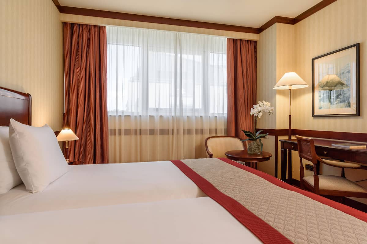 Une chambre d’hôtel avec un grand lit, un bureau, une chaise, une lampe et une fenêtre avec des rideaux.