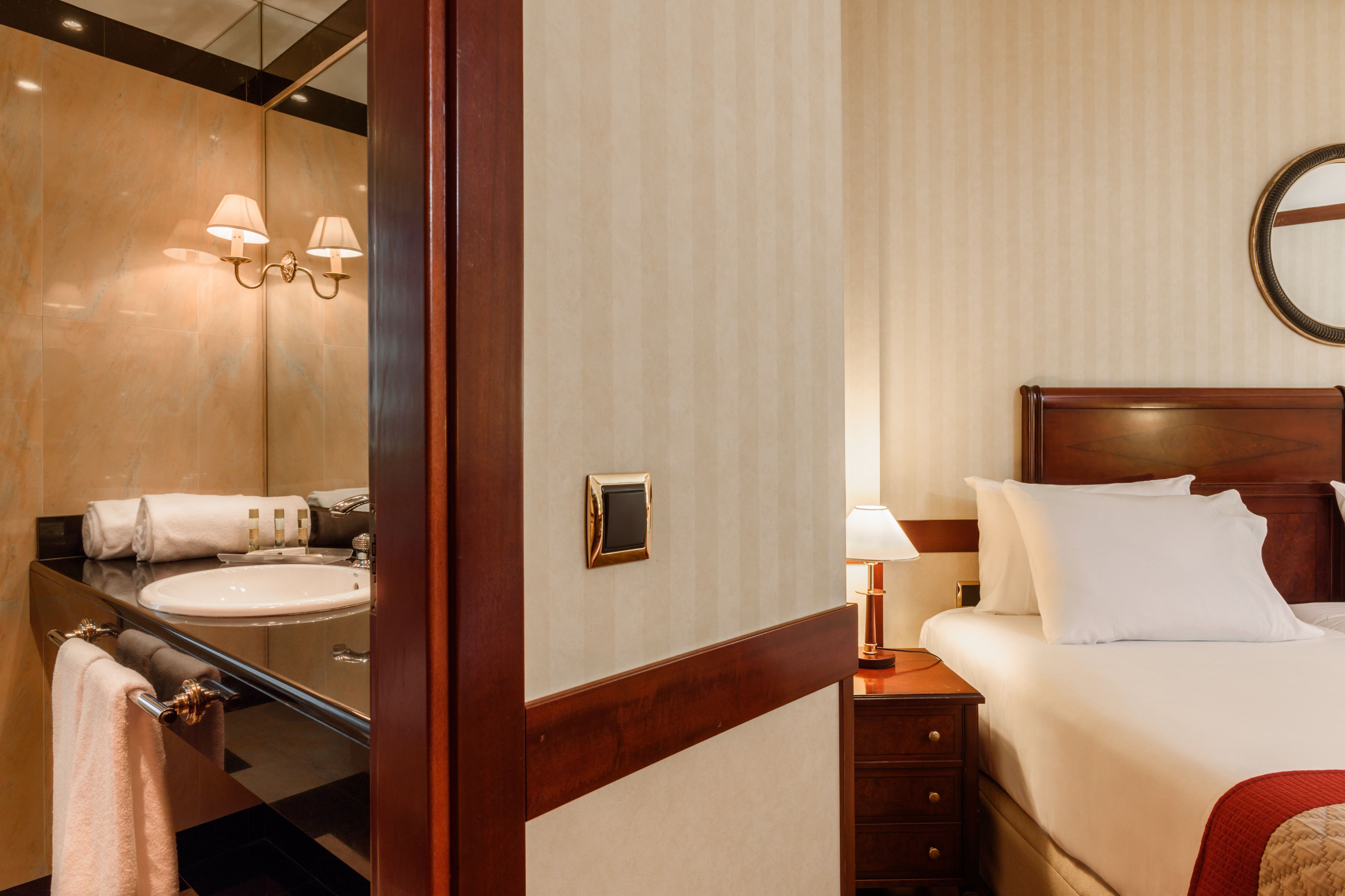 A hotel room with a wooden headboard, a large mirror, a bedside table with a lamp, and a bed with white linens.