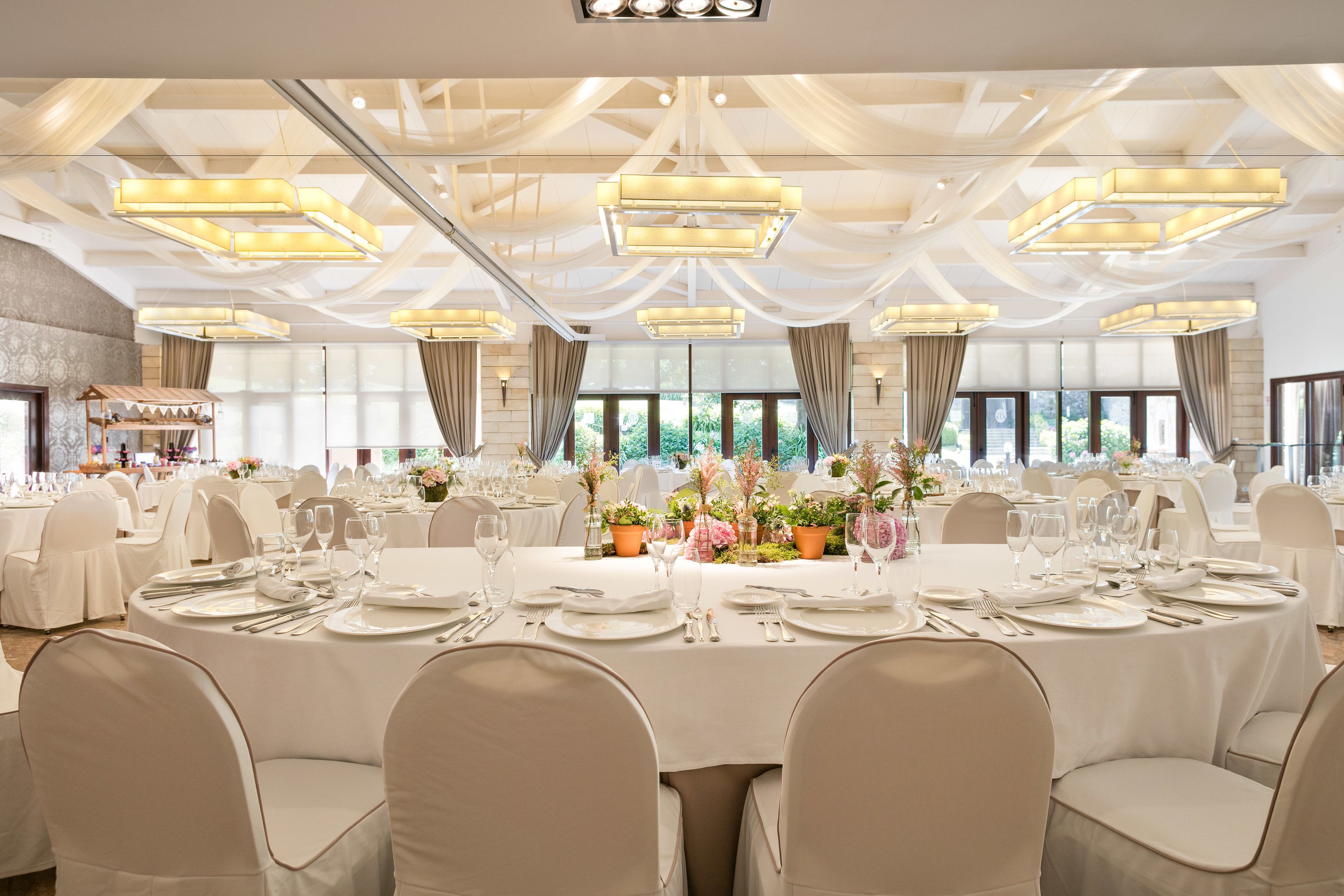 A banquet hall with round tables set for dining, large chandeliers, and a view of greenery through the windows.