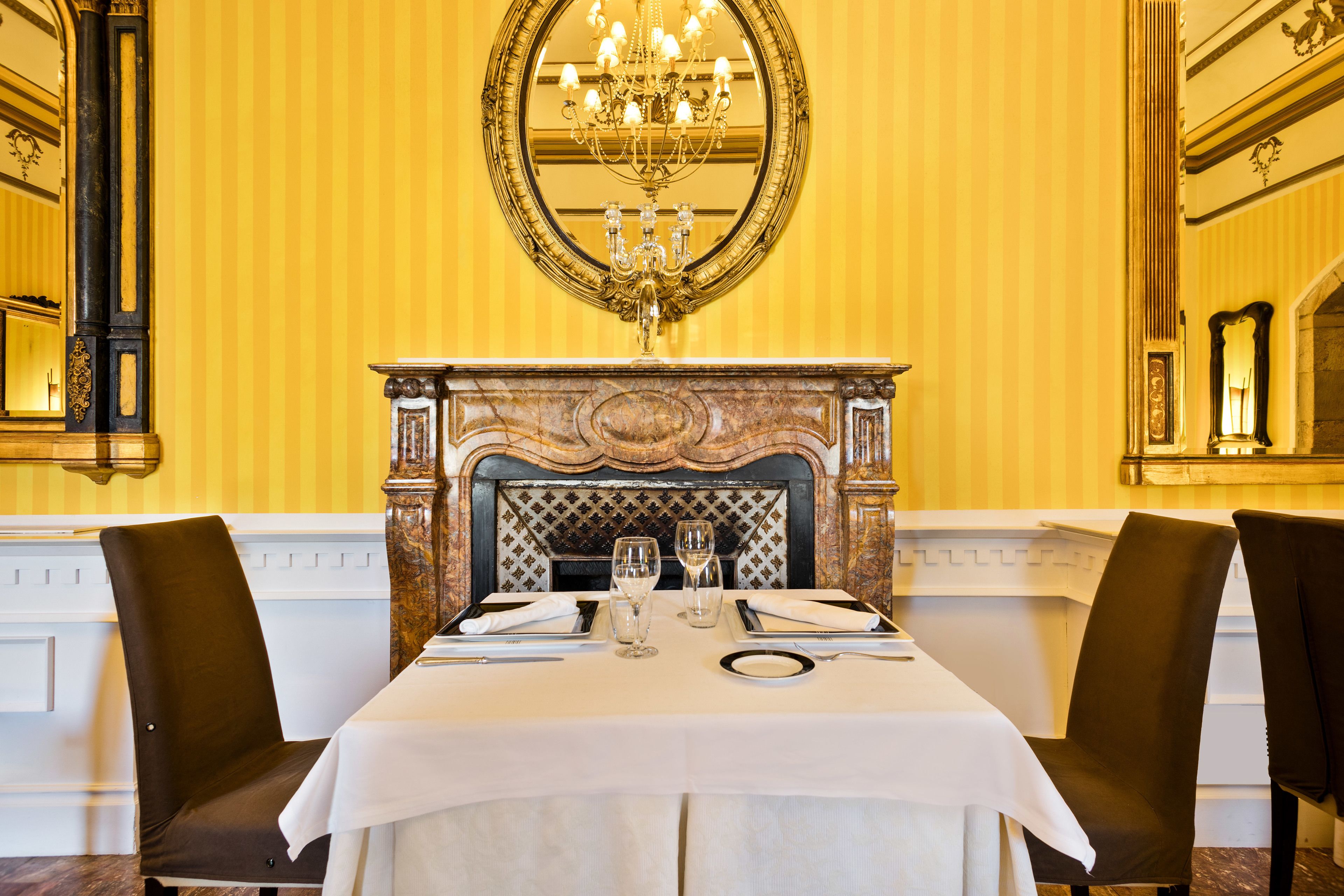 Un comedor formal con una mesa dispuesta para dos personas, chimenea y decoraciones ornamentales en las paredes.