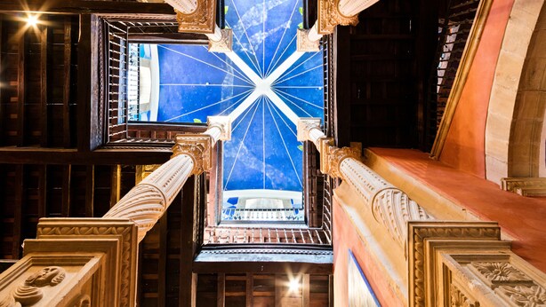 A grand architectural interior with ornate columns, a central skylight, and a view of the sky through a patterned window.