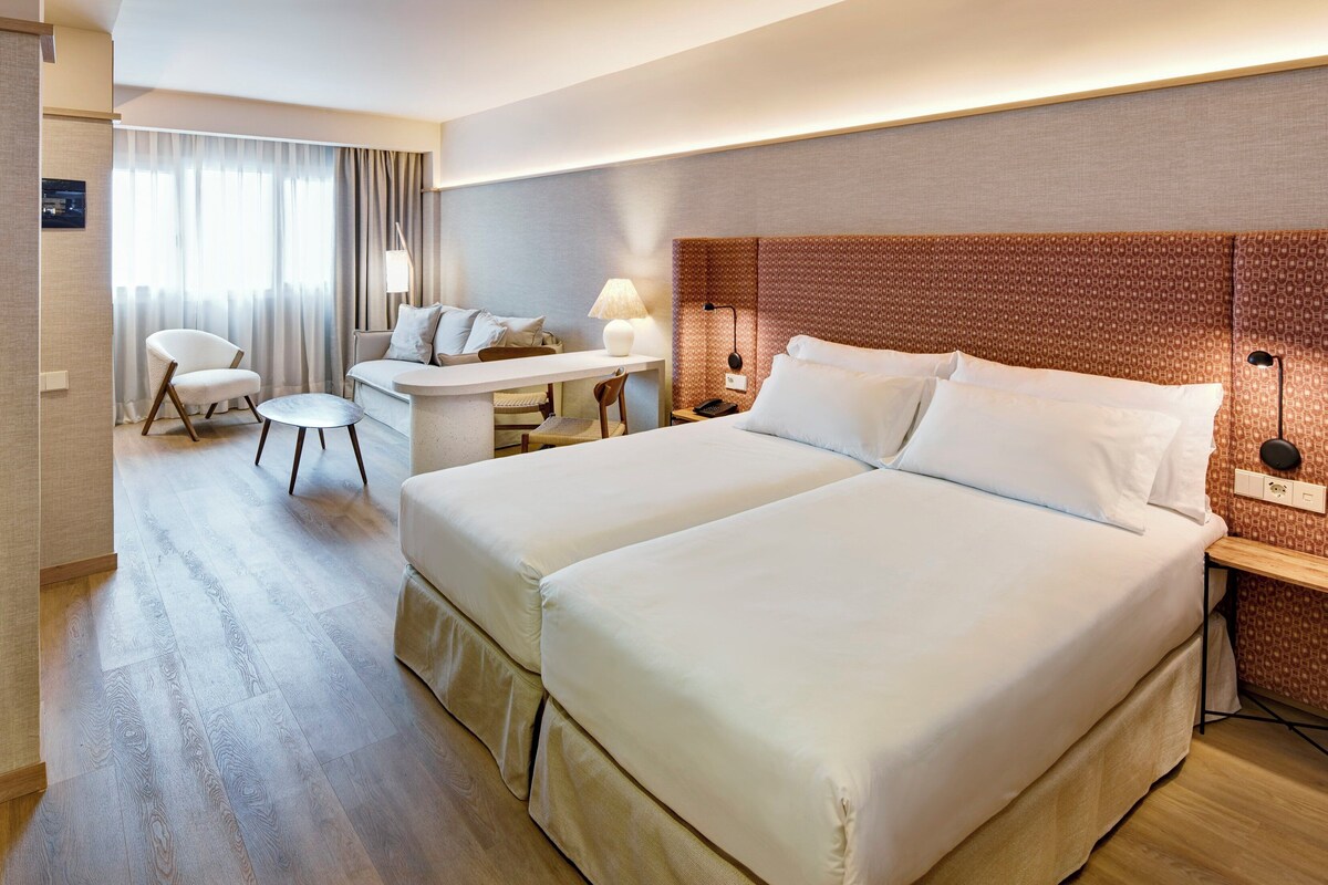 A hotel room with a bed, a desk, a chair, and a lamp.