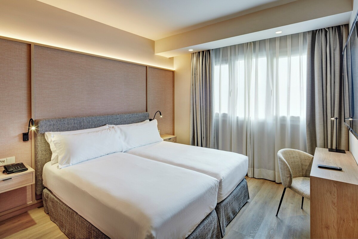 A hotel room with a double bed, bedside tables, a desk with a chair, a TV, and a window with curtains.