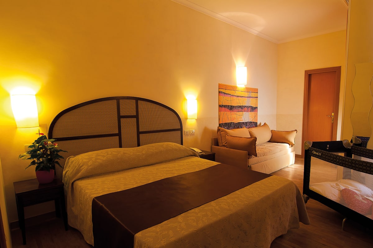 Hotelkamer met een bed, nachtkastje met een lamp, een stoel, een klein tafeltje en een schilderij aan de muur.