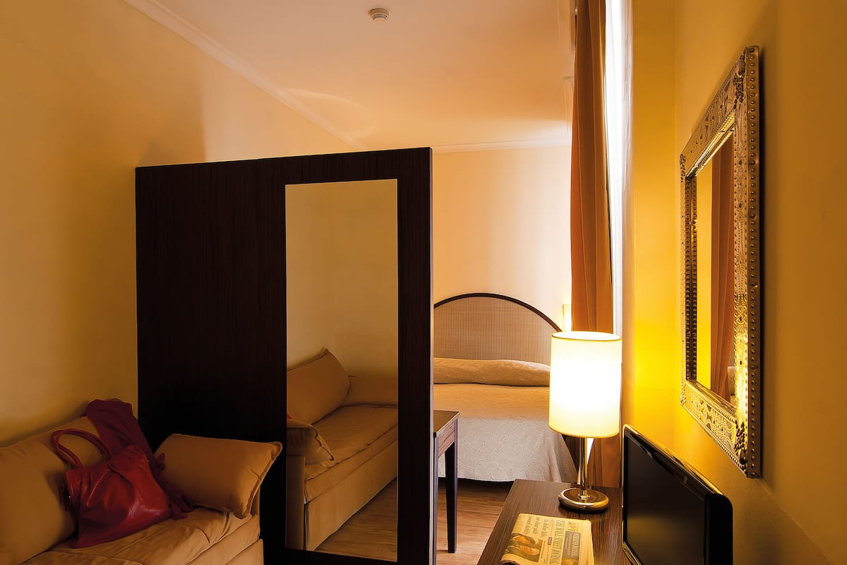 Een hotelkamer met een bed, een nachtkastje met een lamp, een spiegel en een rode handtas op de bank.