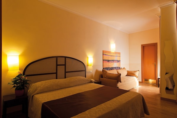 Una habitación de hotel con una cama grande, mesitas de noche, una pequeña mesa, una silla, una planta en maceta y un cuadro en la pared.