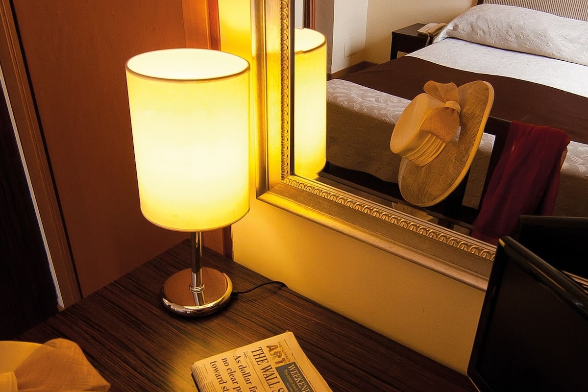 Hotelkamer met een bedlampje, een krant, een hoed op het bed en een spiegel die de kamer weerspiegelt.