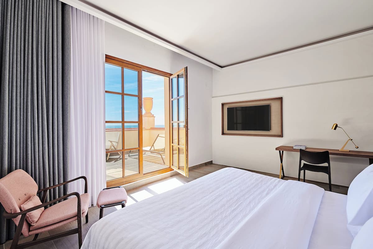 A hotel room with a large bed, a desk, a chair, and a balcony with a view of the sea.