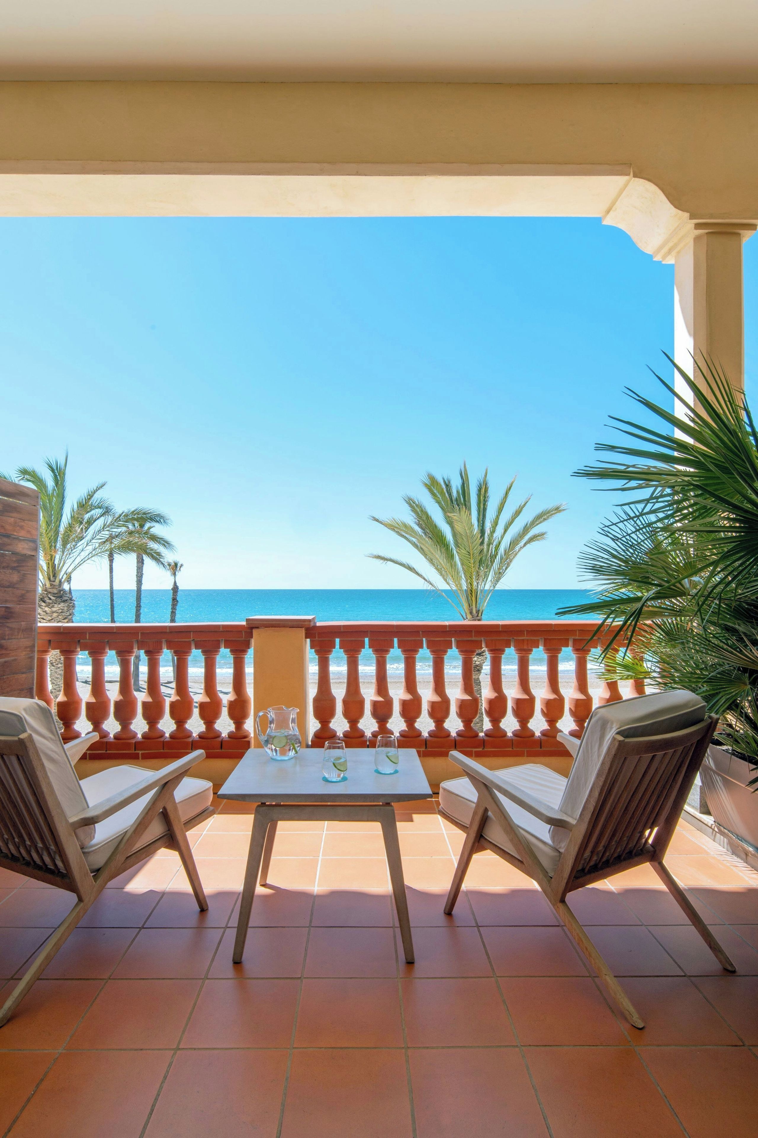 A balcony with two chairs and a table, overlooking a beach with palm trees and the sea.