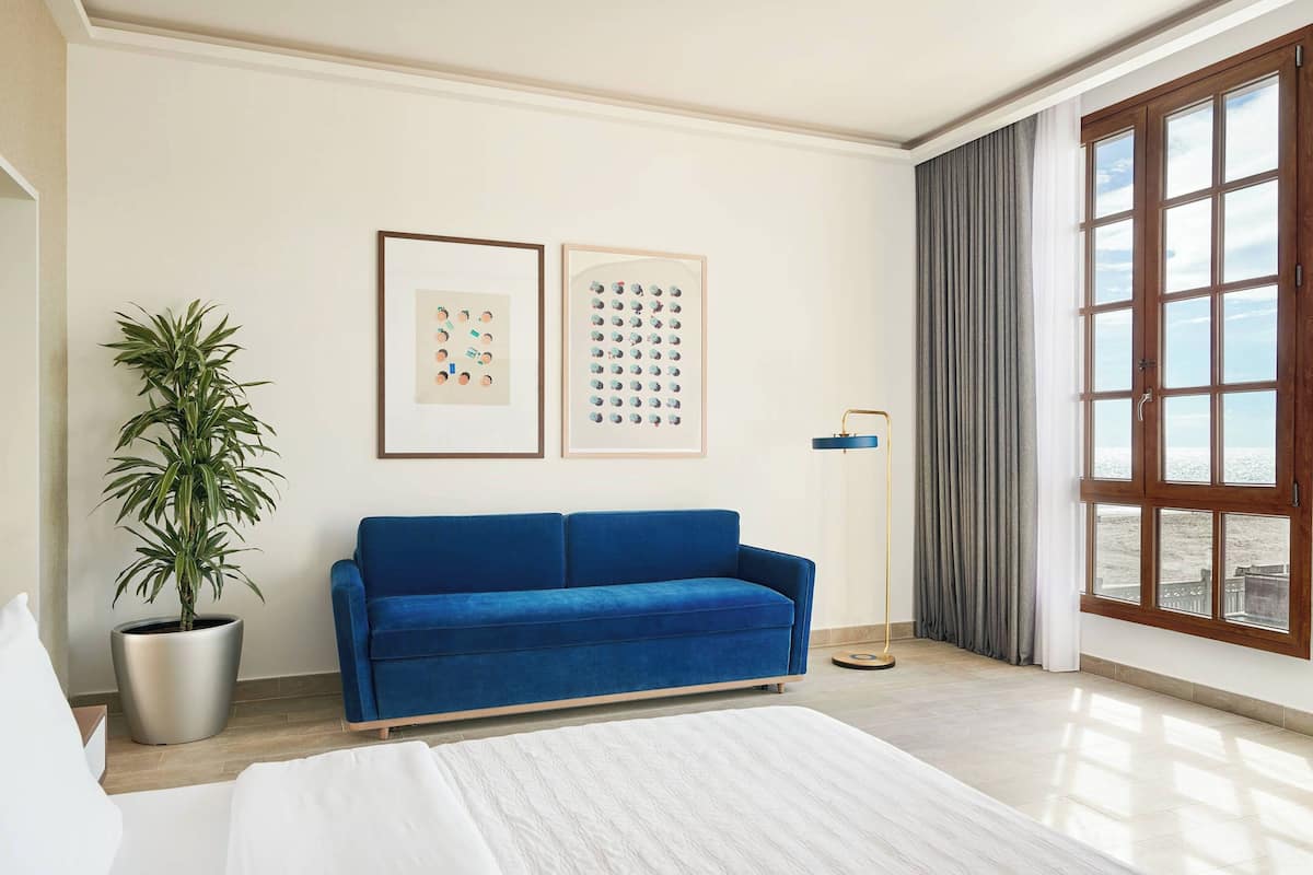 A modern hotel room with a large bed, a blue sofa, a potted plant, and a large window with a view of the beach.