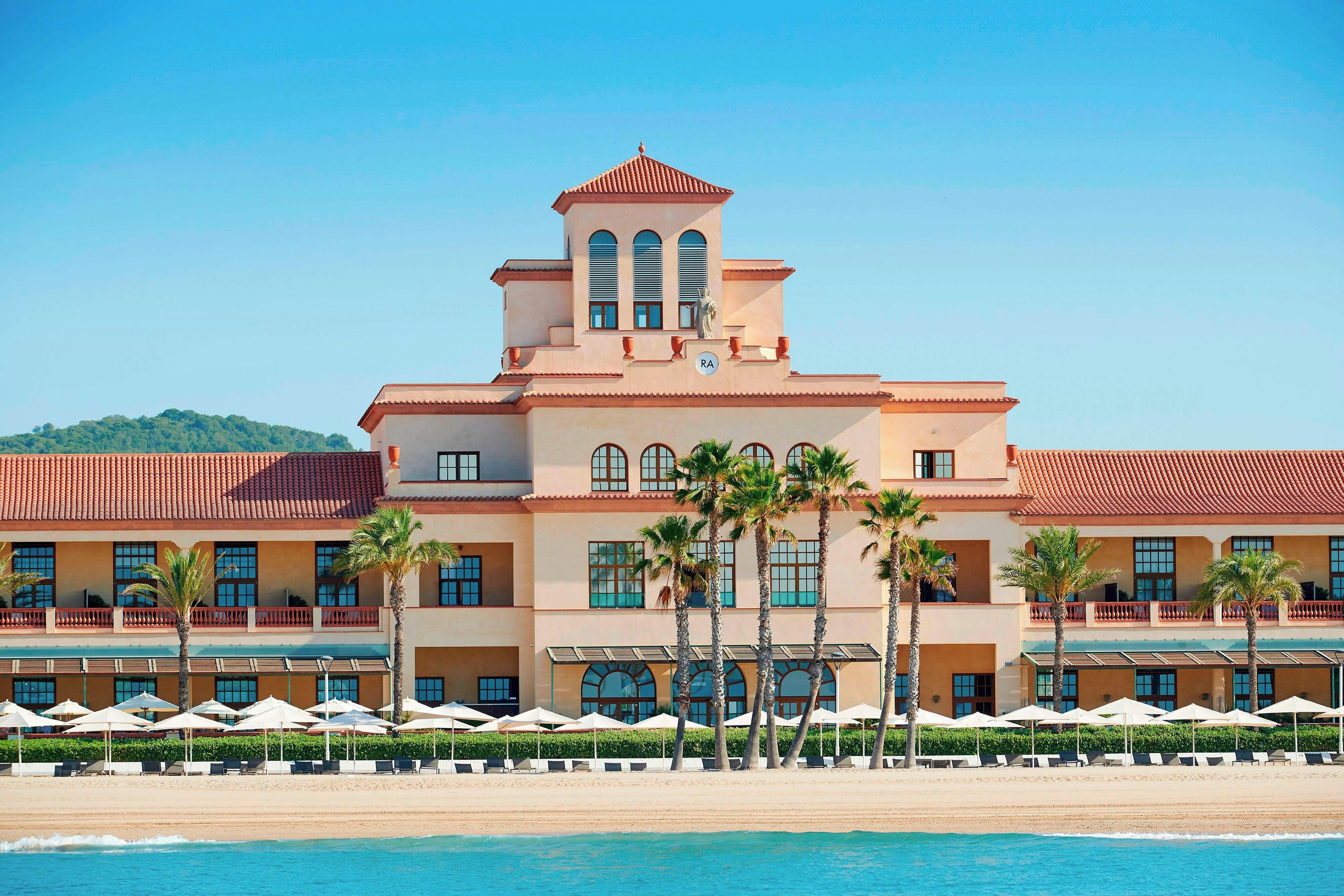 A hotel with a poolside area, palm trees, and a clear blue sky.