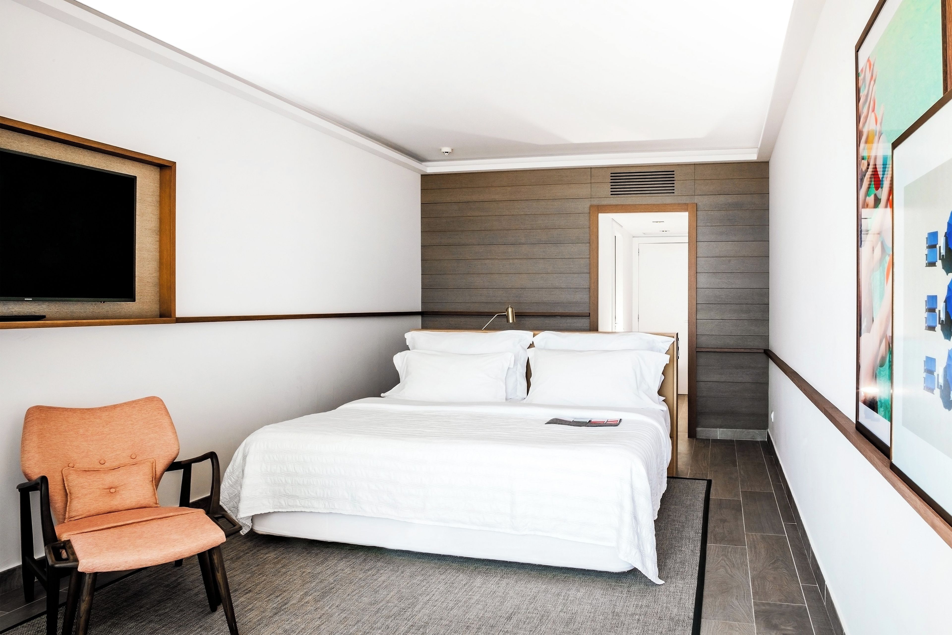 A hotel room with a bed, a chair, a TV, and a door leading to another room.