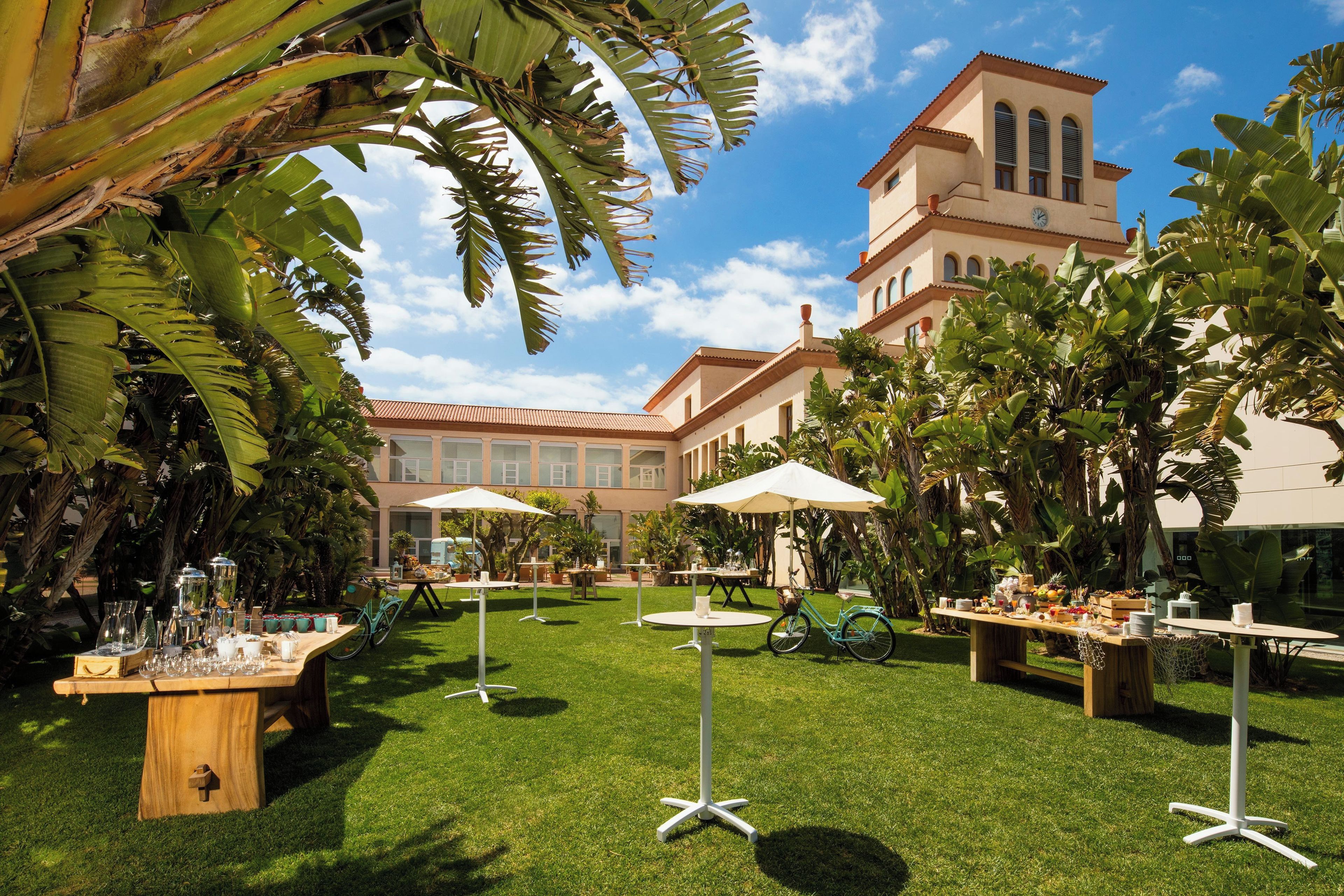 An outdoor dining area with tables and umbrellas, surrounded by lush greenery and a building with arched windows.