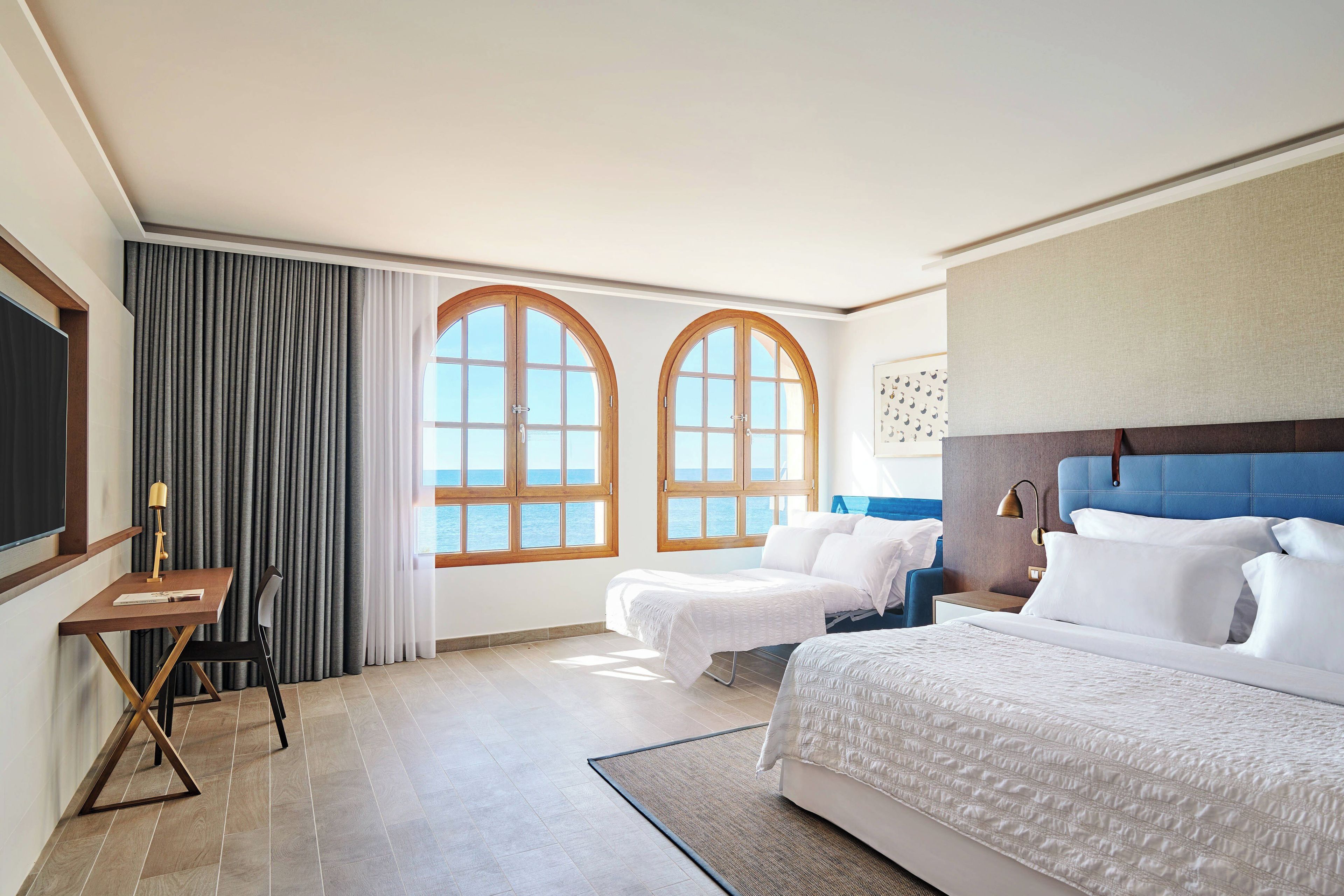 A hotel room with a large bed, a desk with a chair, two windows, and a view of the sea.