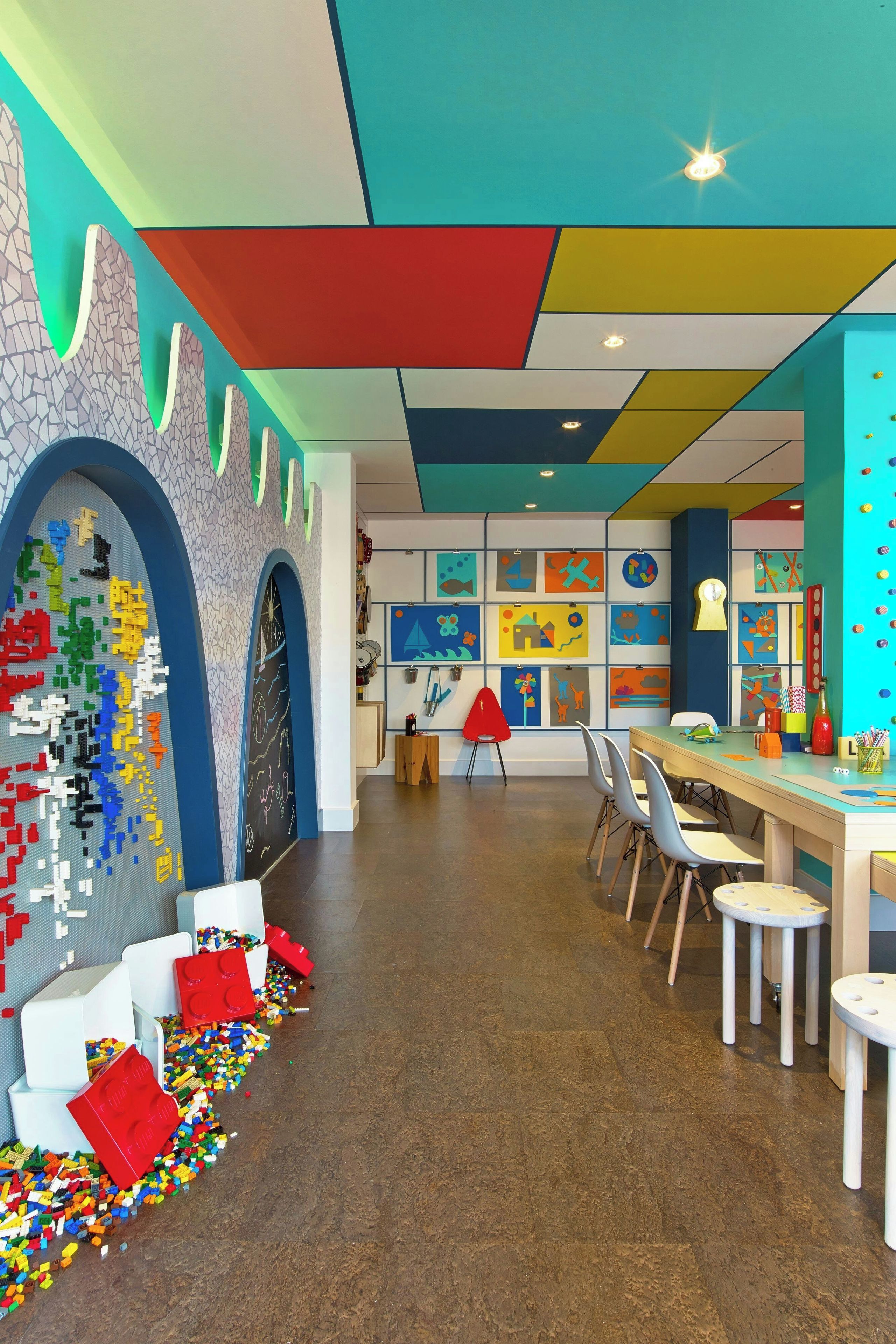 Un área de juegos infantil colorida con paredes de LEGO, una mesa, sillas y una variedad de juguetes.