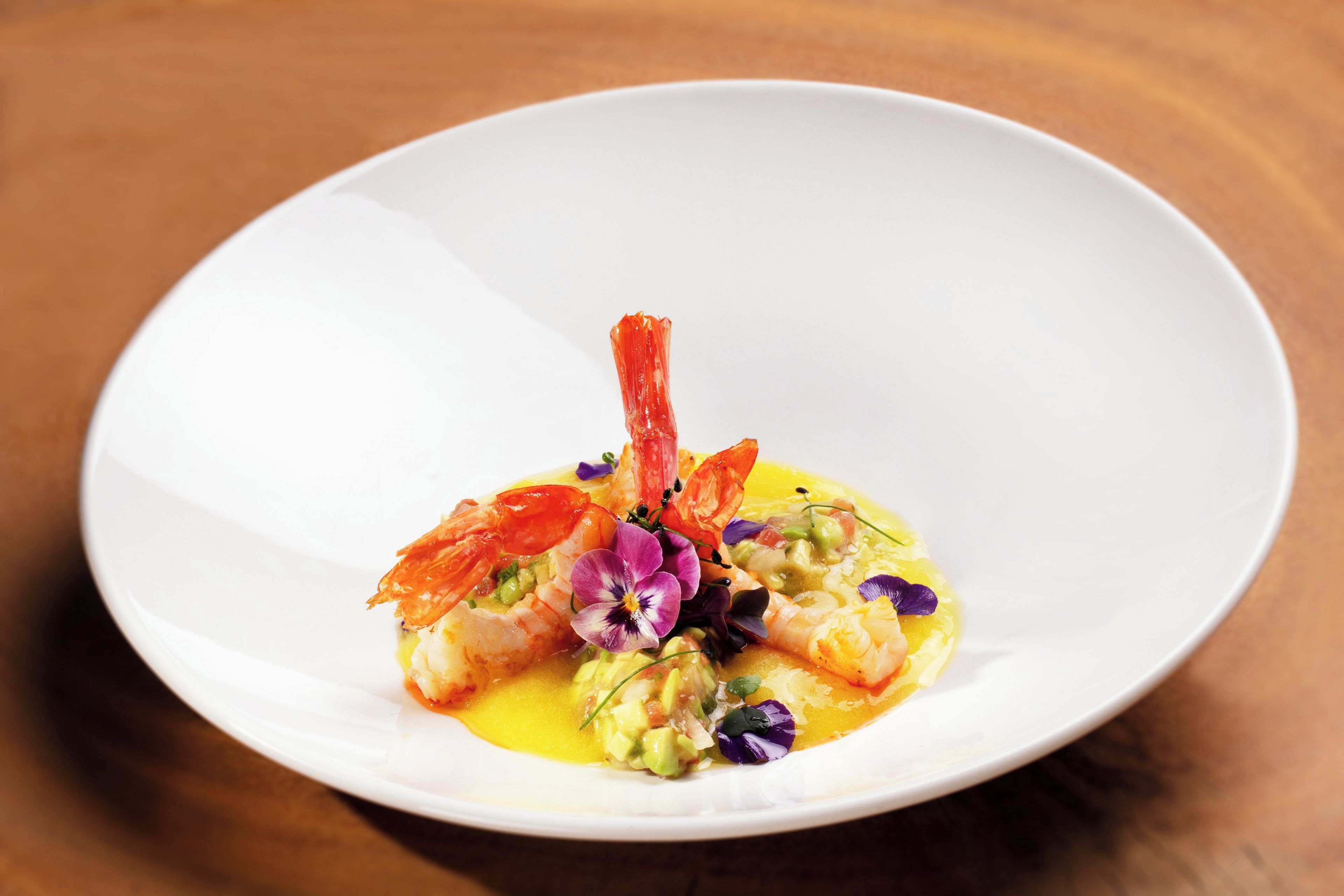 A white plate with a seafood dish, possibly shrimp, garnished with purple flowers and a yellow sauce.