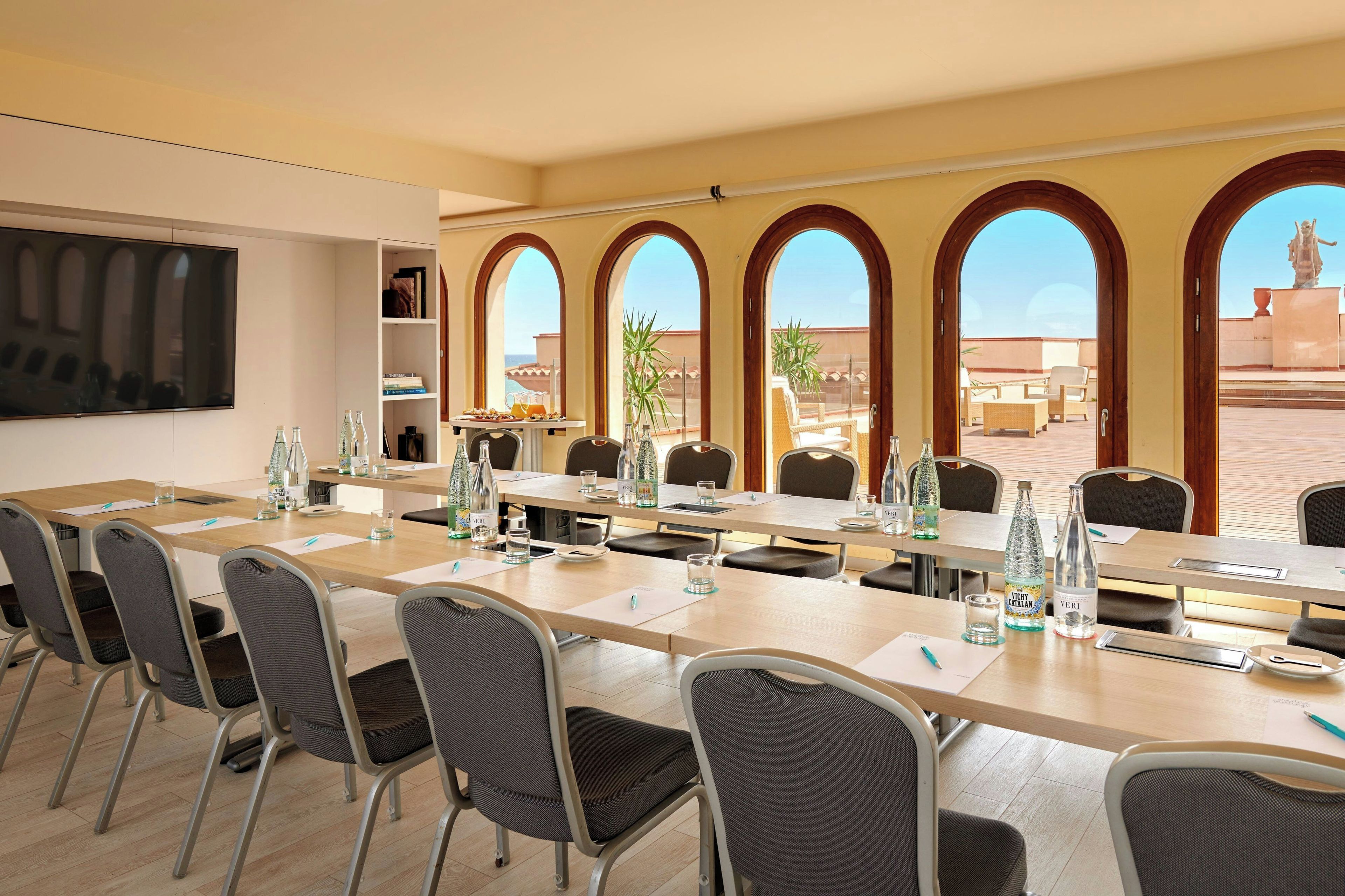 A conference room with a long table, chairs, a TV, and a view of the outside through arched windows.