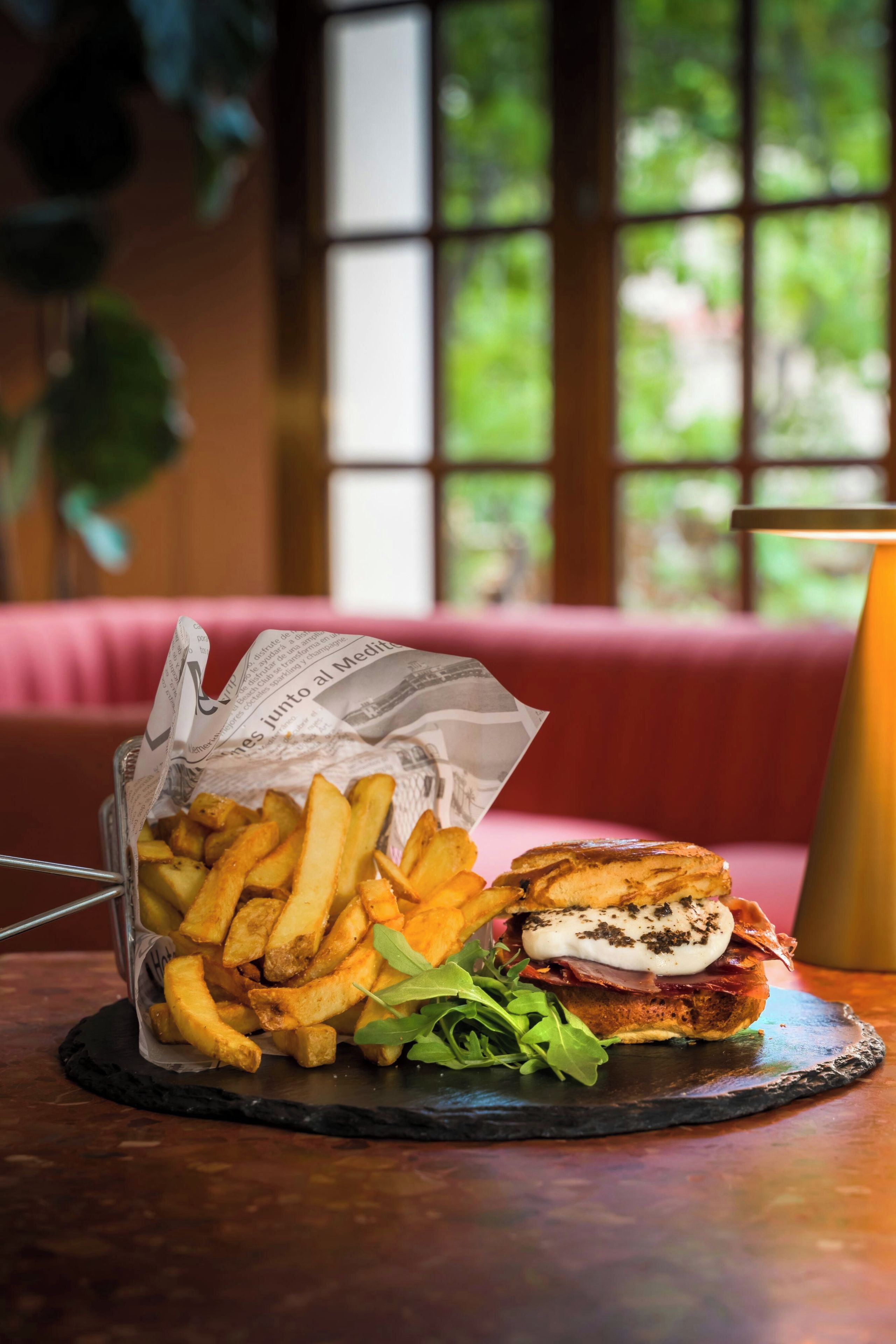 A sandwich with fries on a slate plate, a drink in a glass, and a red cushioned bench in the background.