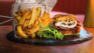 A sandwich with fries on a slate plate, a drink in a glass, and a red cushioned bench in the background.
